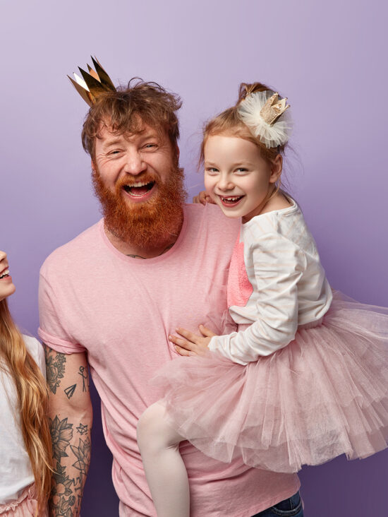 Vertical shot of overemotive joyful single dad and two daughters, celebrate Fathers Day, wear festive outfits, have fun together, isolated over purple background, blank space above for promotion
